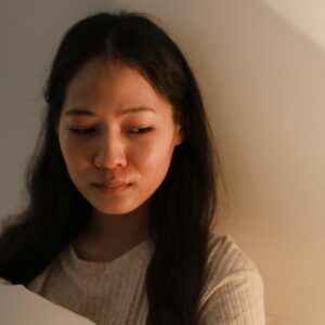 A young woman sitting indoors with a melancholic expression, dim lighting.